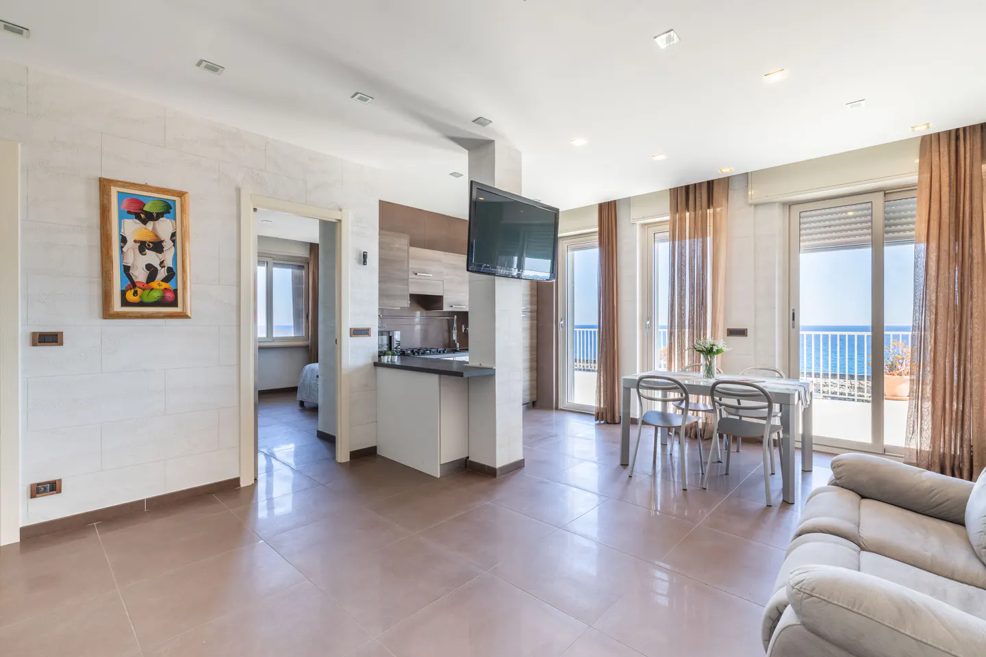 Bright, open apartment with a kitchen, dining table, and ocean view. A painting hangs on the wall.