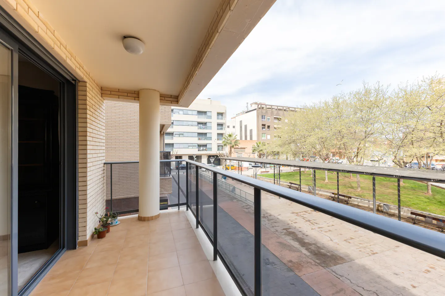 A bright balcony with tan tile flooring and a glass railing overlooks a park with trees and benches. A sliding glass door leads inside.