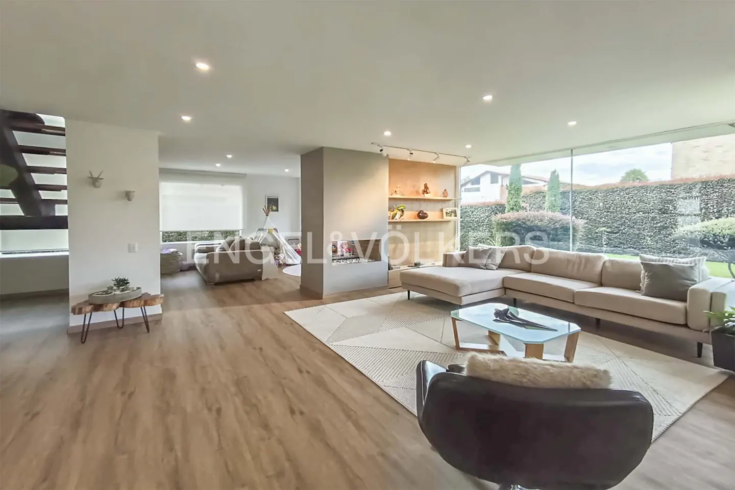 Modern open-plan living room with light wood floors, a large beige sectional, glass coffee table, and floor-to-ceiling glass.