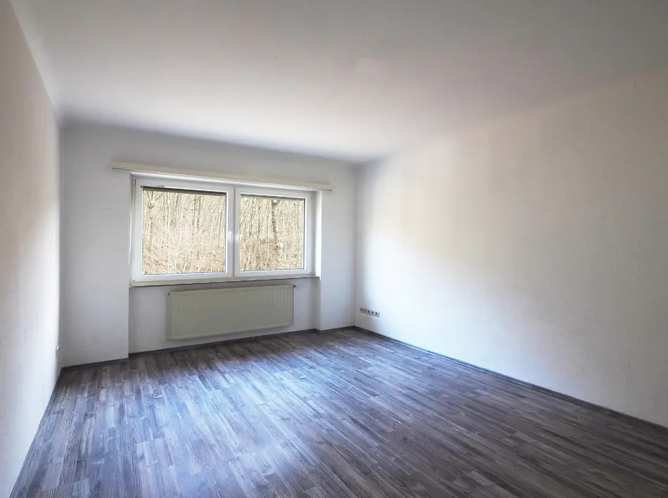 Bright, empty room with gray wood floors, white walls, and a window overlooking a forest. A radiator sits beneath the window.