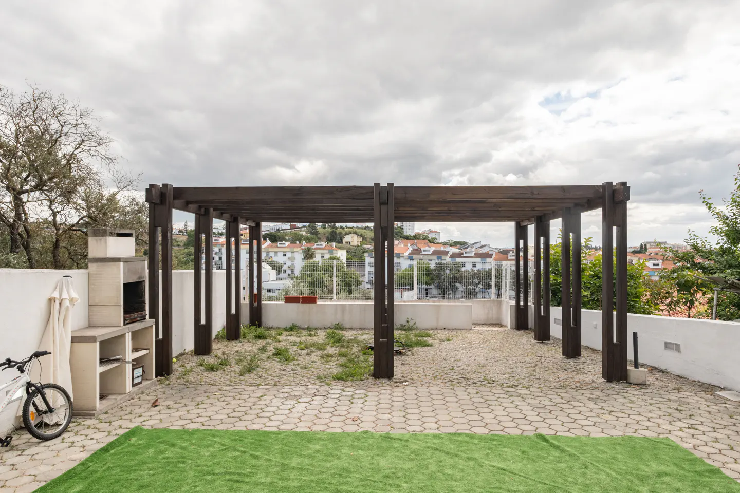 Outdoor patio with a dark wood pergola, stone pavers, and a green rug. A white grill and bicycle are on the left. Cityscape in the background.