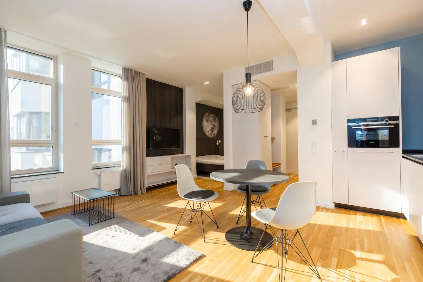 Bright, modern apartment interior with wood floors, white walls, and large windows. A round table with chairs sits under a pendant light.
