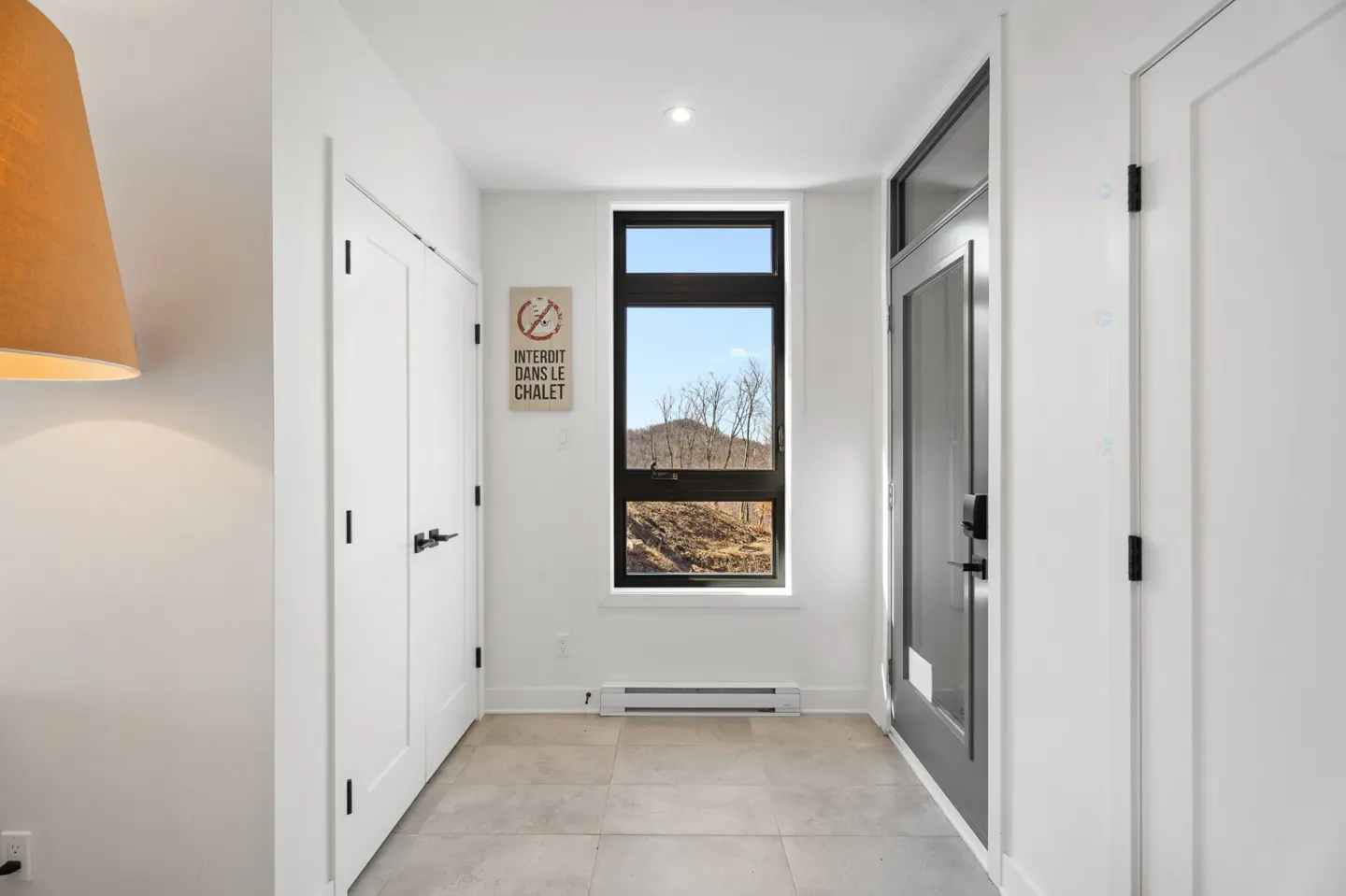Hallway with white walls, tile floor, and black door hardware. A window shows a mountain view. A sign reads "Interdit dans le chalet."
