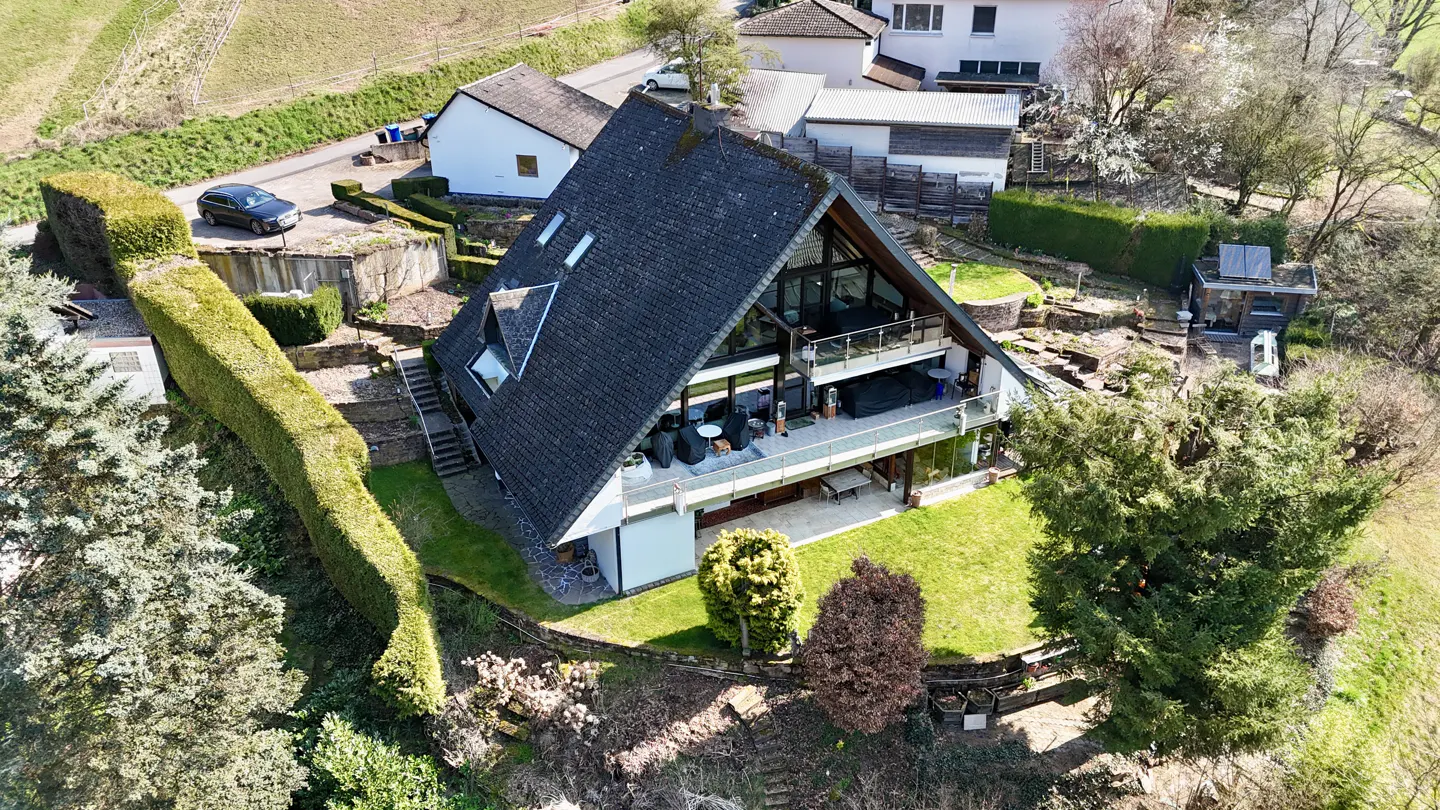 Aerial view of a modern A-frame house with black roof, glass walls, and balconies, surrounded by green lawns and trees.