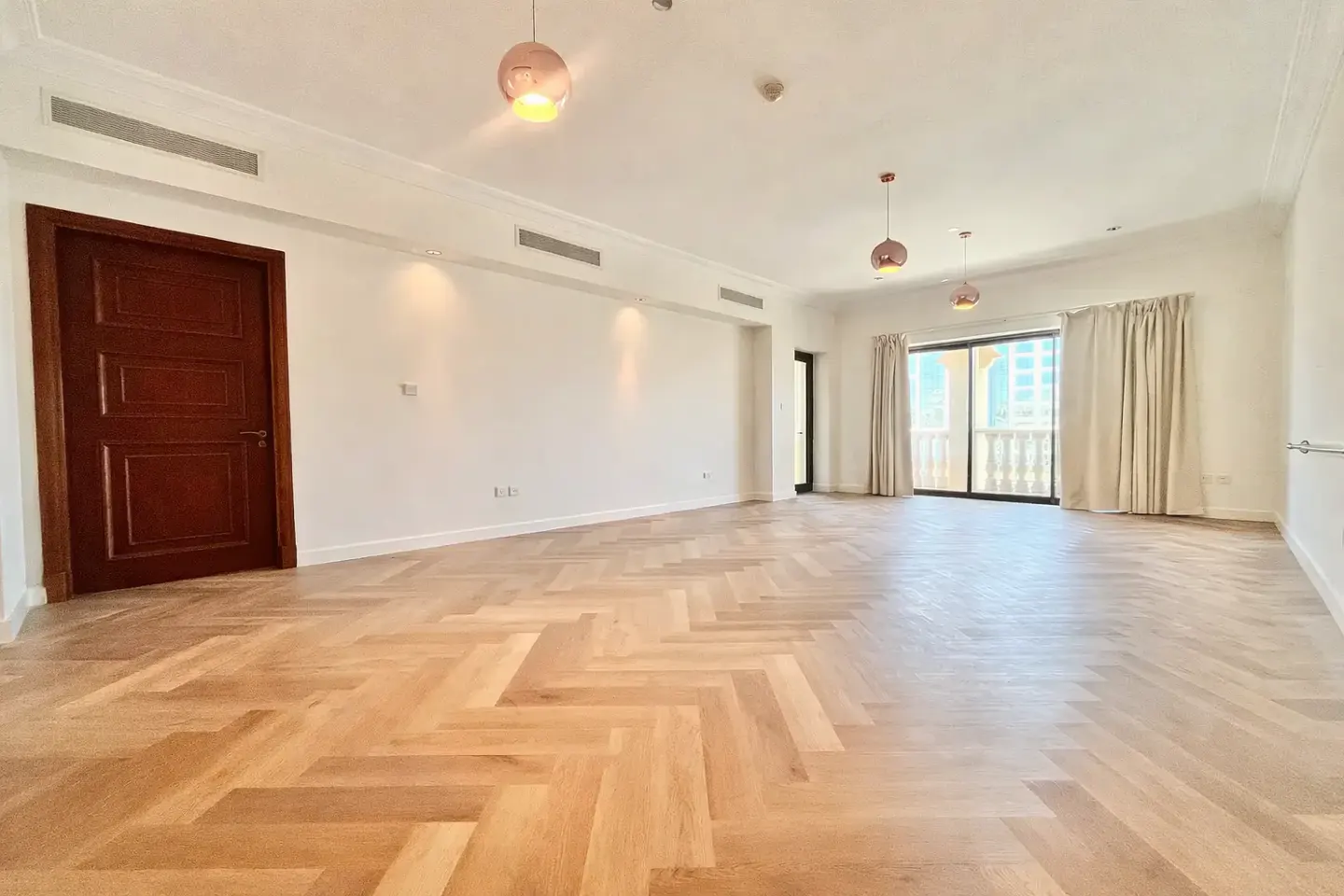 Bright, empty room with herringbone wood floors, white walls, and a dark wood door. Sliding glass doors lead to a balcony.