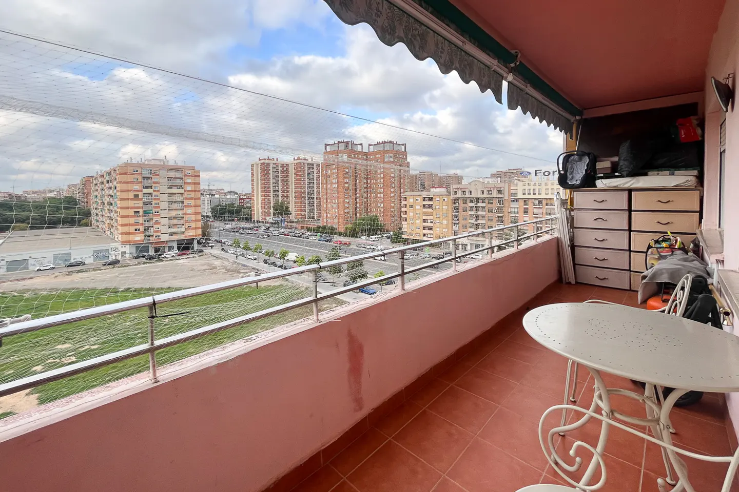 Balcony view with white metal table and chairs, pink walls, and a city view with tall buildings and a cloudy sky.