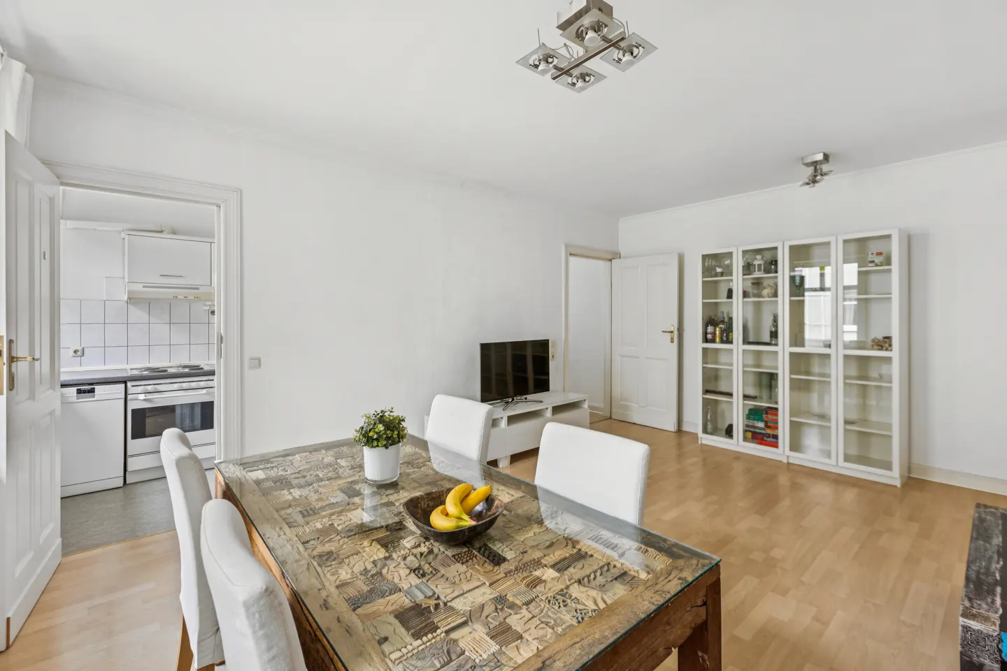 Bright dining room with a patterned wood table, white chairs, and a bowl of bananas. Kitchen visible through doorway.