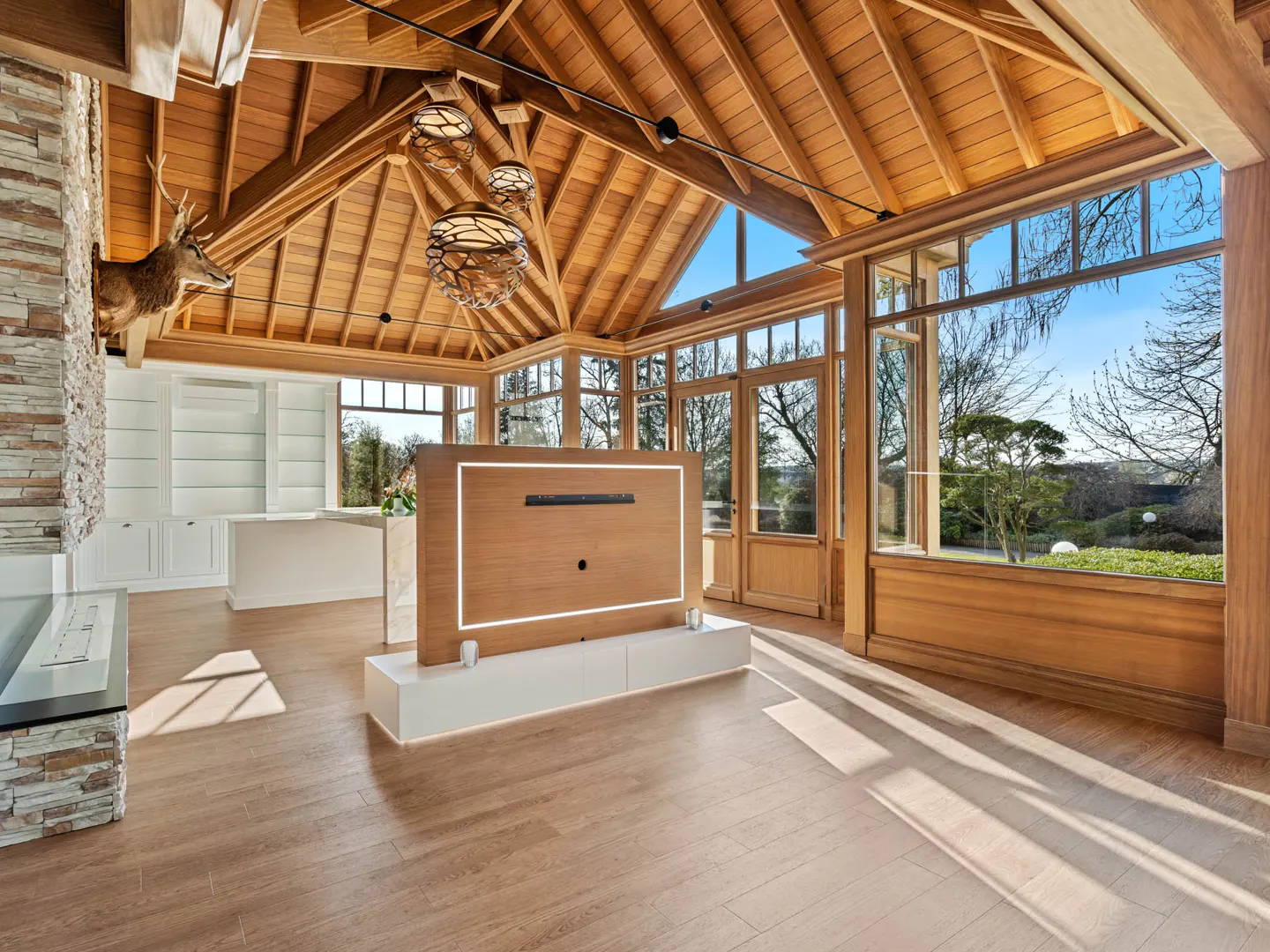 A sunroom with wood floors, a vaulted wood ceiling, and large windows overlooking a garden. A deer head is mounted above a stone fireplace.