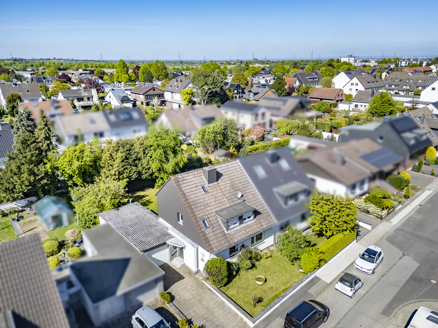 Aerial view of a gray and white house with a tile roof, green lawn, and cars parked on the street in a suburban neighborhood.