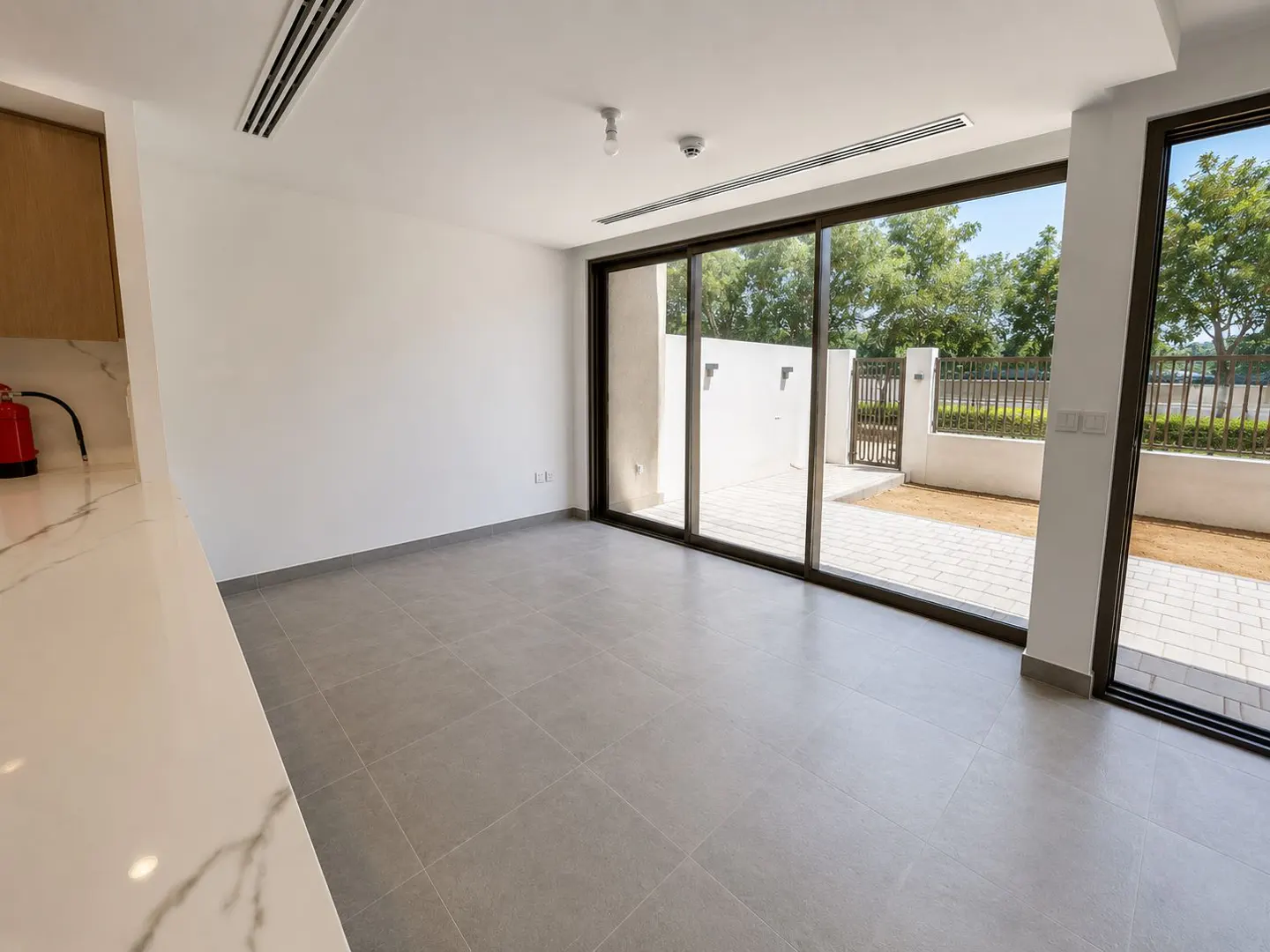 An empty room with gray tile floors and white walls. Large sliding glass doors lead to a patio and backyard. A white marble countertop is visible on the left.