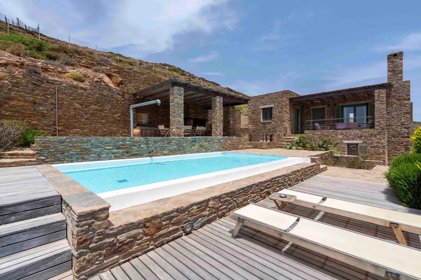 Stone villa with a swimming pool, wooden deck, lounge chairs, and a covered patio against a rocky hillside.