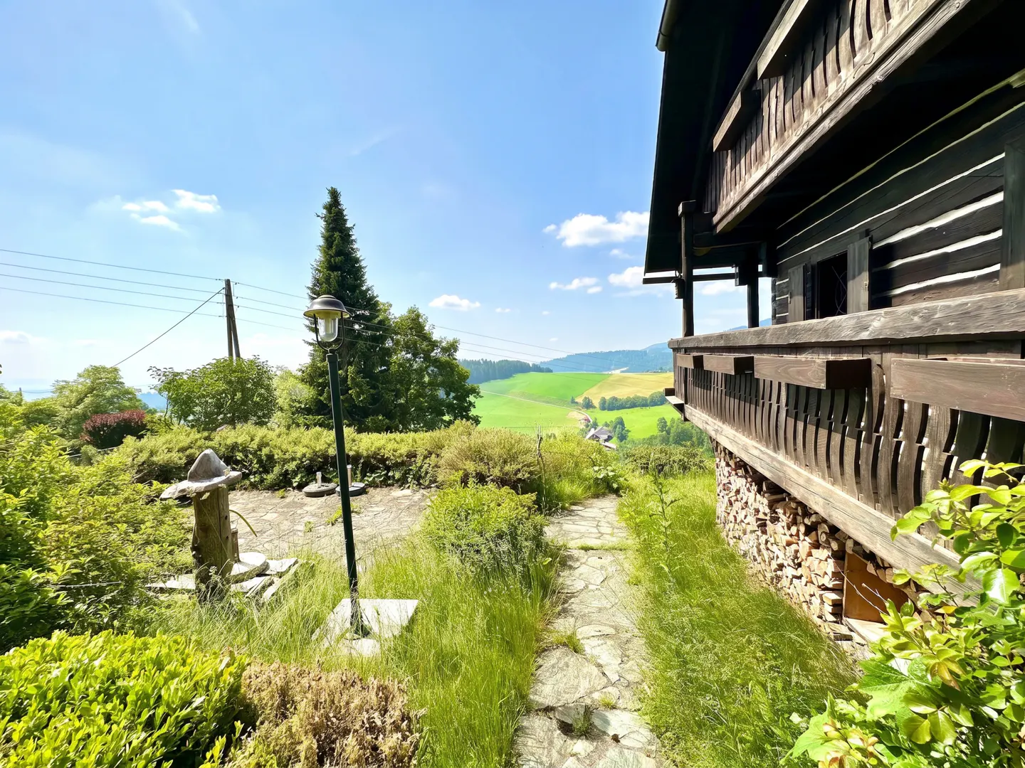 Exterior view of a dark wood cabin with a balcony overlooking a green valley under a blue sky. A stone path leads to the cabin.