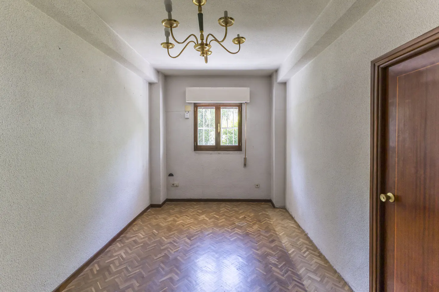 Empty room with white walls, herringbone wood floors, a central brass chandelier, and a window with a wooden frame.