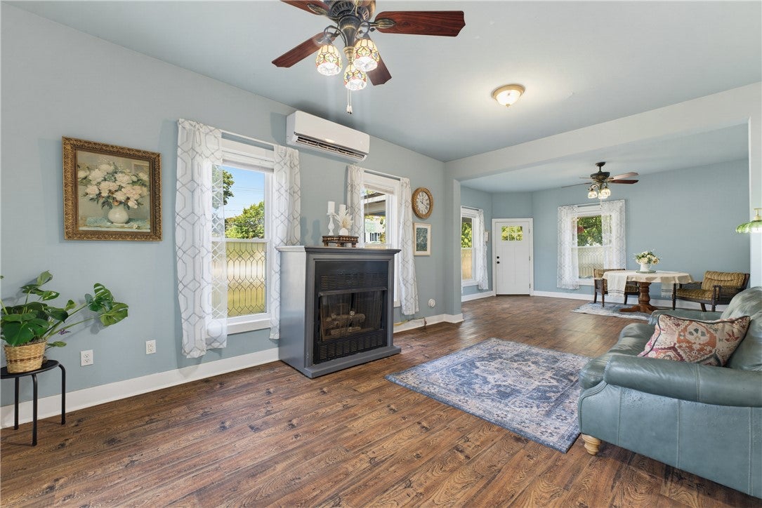 Light blue living room with hardwood floors, a fireplace, ceiling fan, and a view into a dining area with a white door. [AI-generated text]