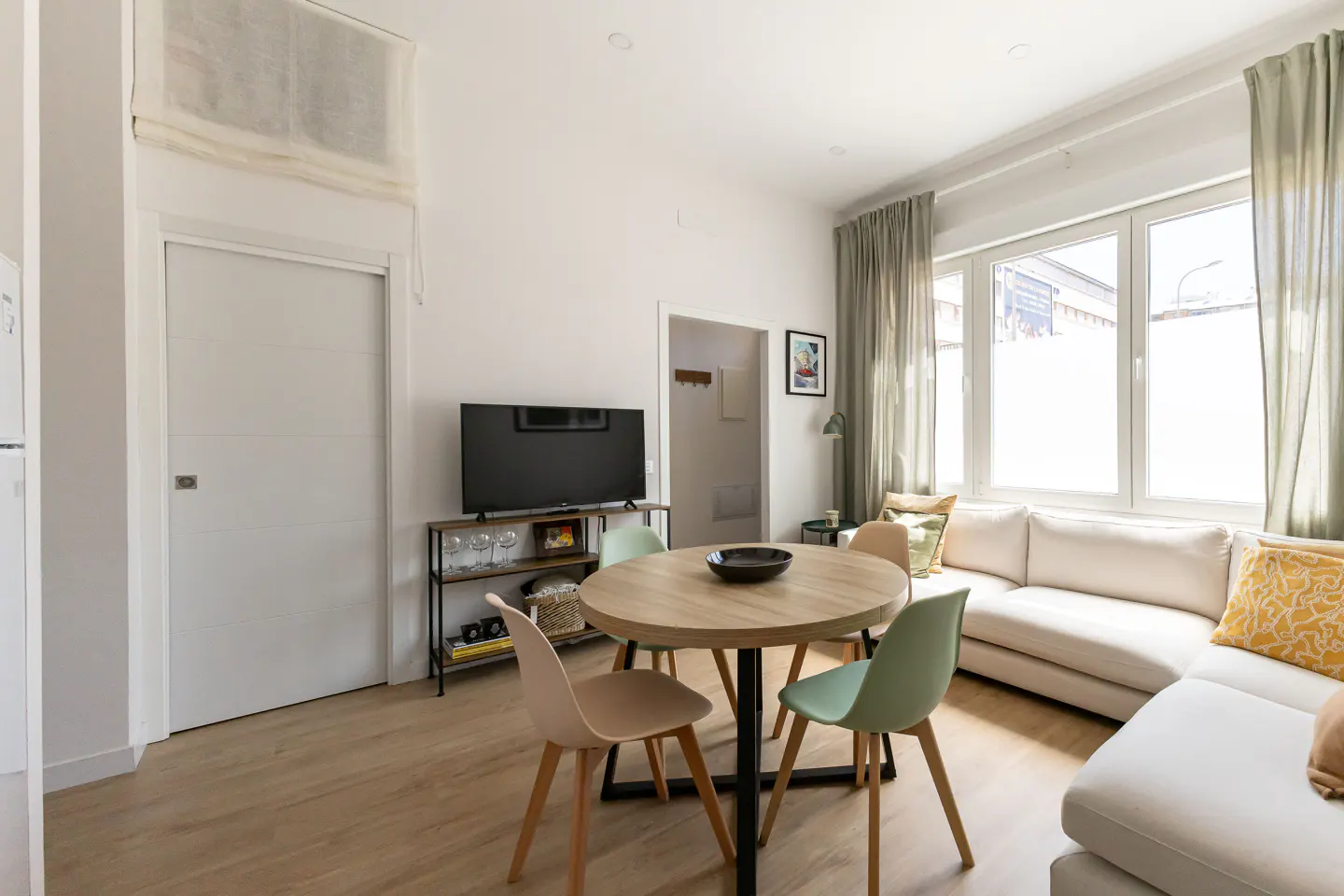 Bright living room with a round wooden table, pastel chairs, and a white sectional sofa by a large window. A TV sits on a shelf.