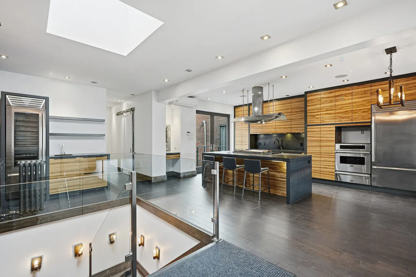 A modern kitchen with wood cabinets, a black countertop island with stools, and stainless steel appliances. A skylight brightens the white ceiling.