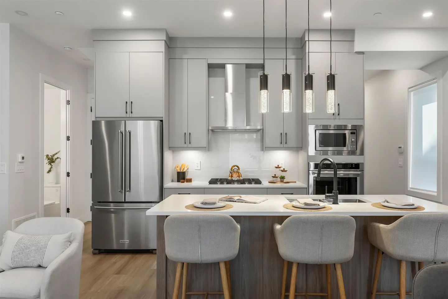 Bright, modern kitchen with gray cabinets, stainless steel appliances, and a white countertop island with four bar stools. Three pendant lights hang above.
