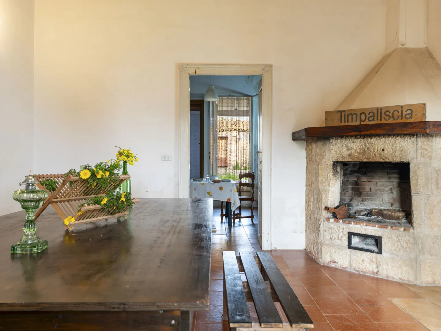 Rustic dining room with a large wooden table, stone fireplace, and a view into another room with a table and chair.