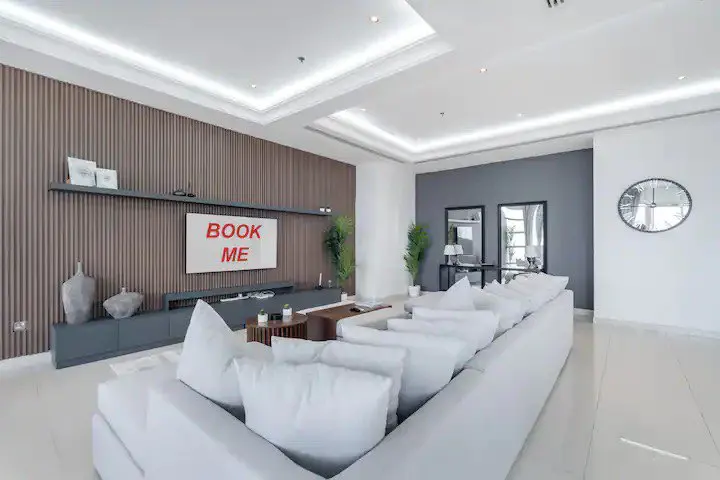 Bright living room with a large white sofa, a TV with "BOOK ME" on the screen, and modern decor.