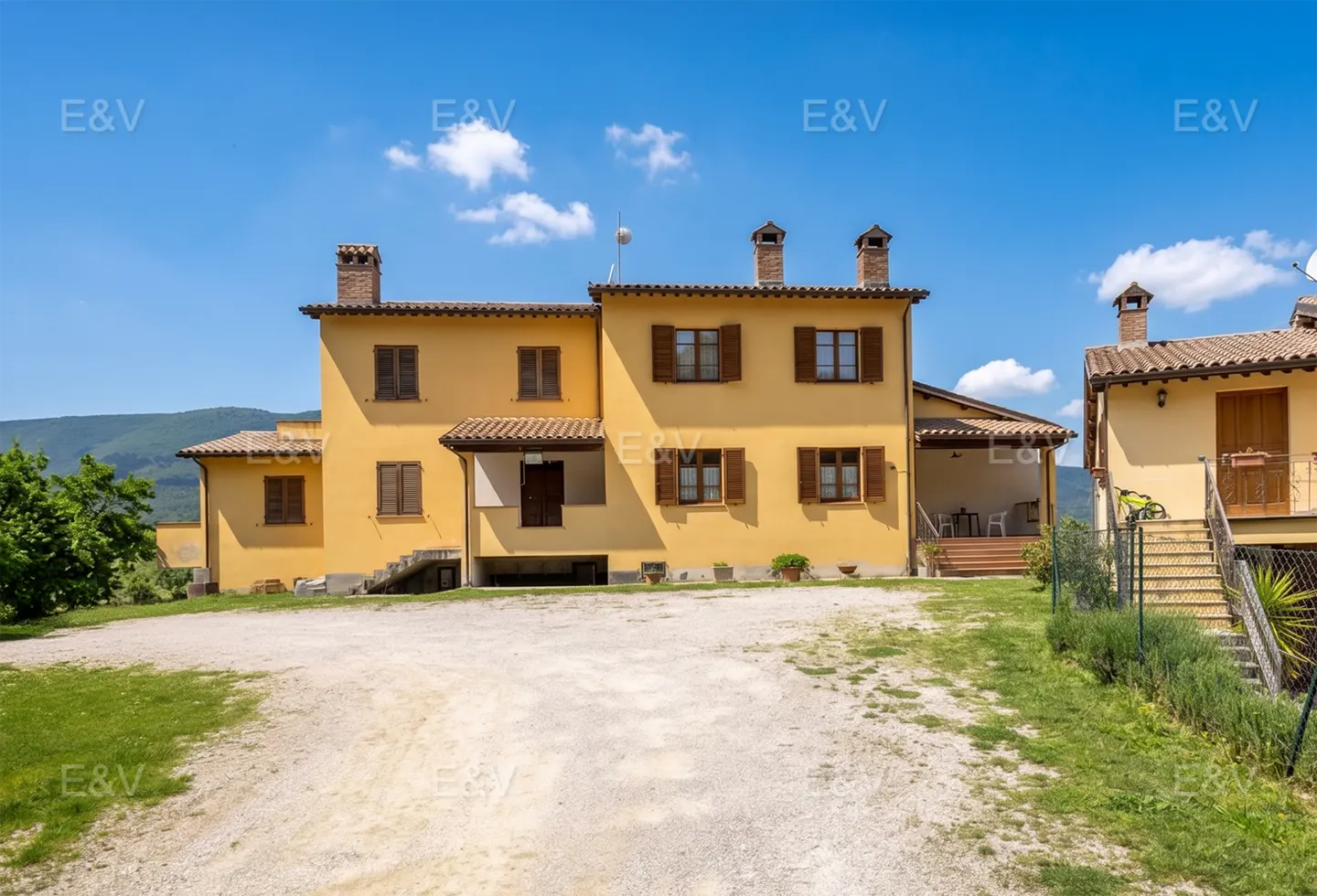 Two-story yellow house with brown shutters and a gravel driveway under a blue sky.