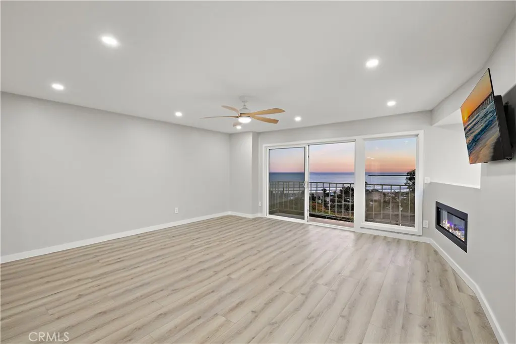 Bright, empty living room with wood floors, ceiling fan, and ocean view through sliding glass doors. A TV and fireplace are on the right wall.