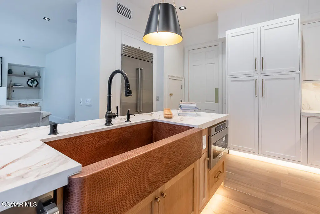 A modern kitchen with a copper farmhouse sink, black faucet, and white marble countertops. White cabinets and stainless steel appliances are visible.