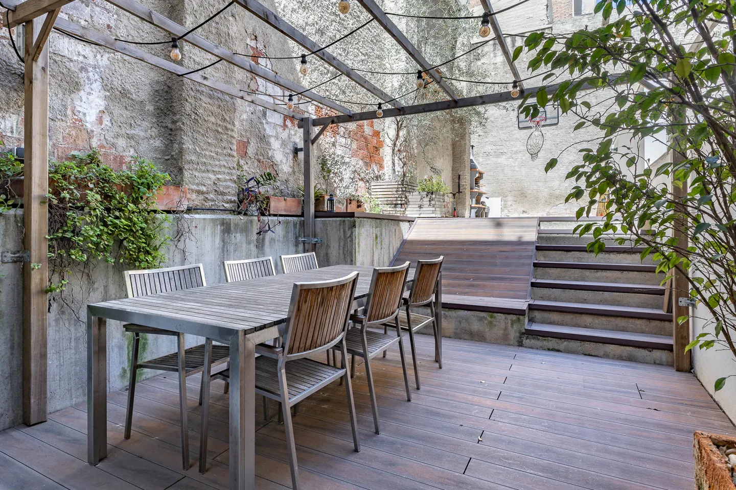 Outdoor patio with wooden table and chairs under a pergola with string lights. Steps lead to an upper level. Brick wall in the background.
