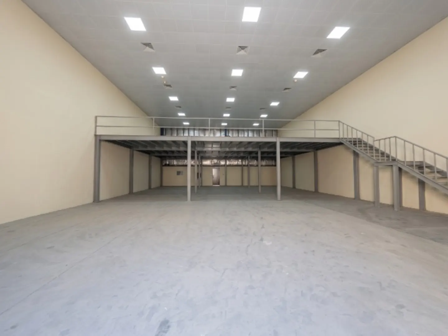 Empty warehouse interior with a gray concrete floor, beige walls, and a metal mezzanine with stairs. Overhead lighting is visible.