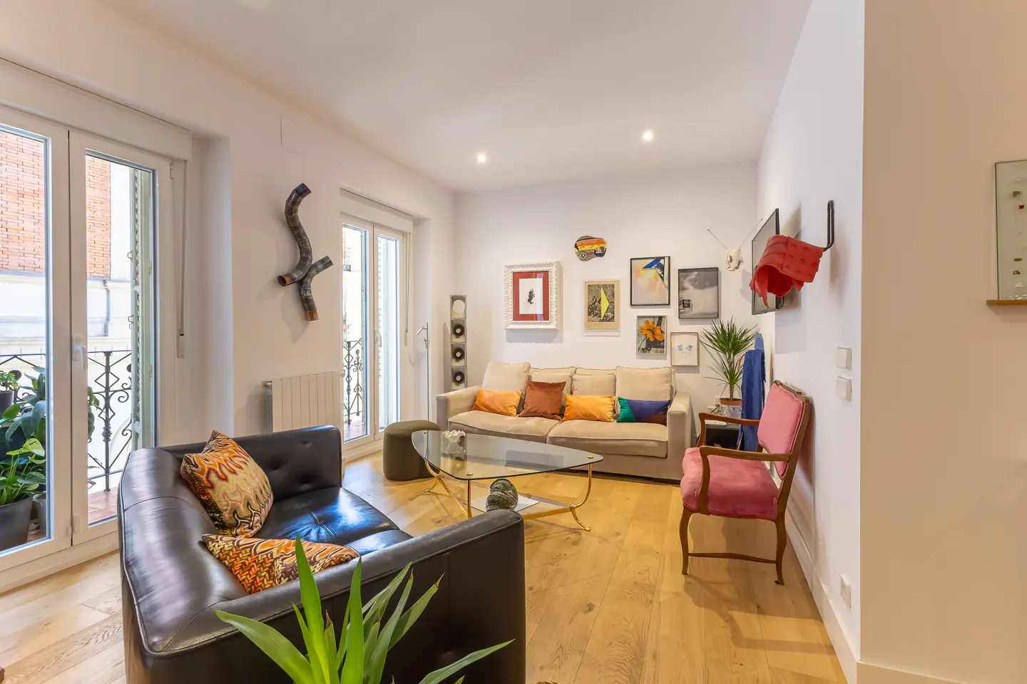 Bright living room with hardwood floors, black leather sofa, beige sofa with colorful pillows, glass coffee table, and gallery wall. Balcony with plants visible.