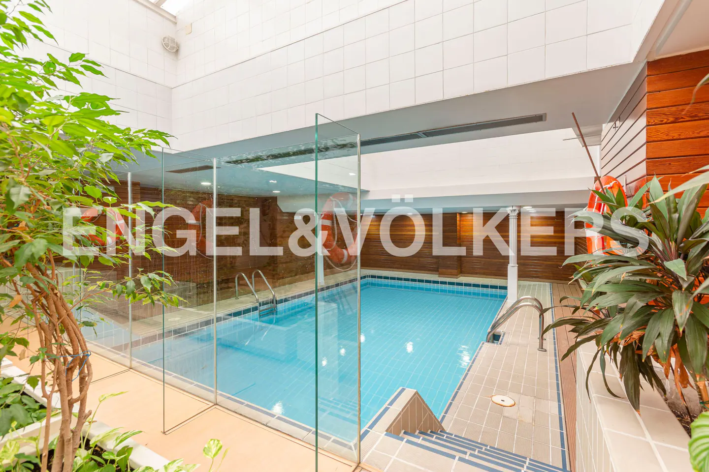 Indoor pool with blue water, surrounded by glass and tile. Plants add greenery to the space. Engel & Volkers logo visible.