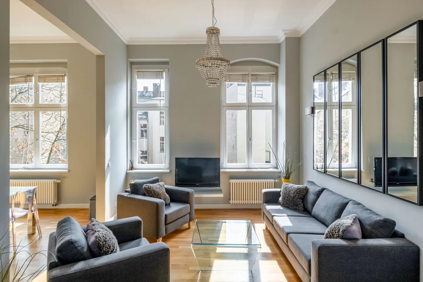 Living room with gray walls, hardwood floors, and large windows. Gray sofa and chairs surround a glass coffee table. A chandelier hangs from the ceiling.