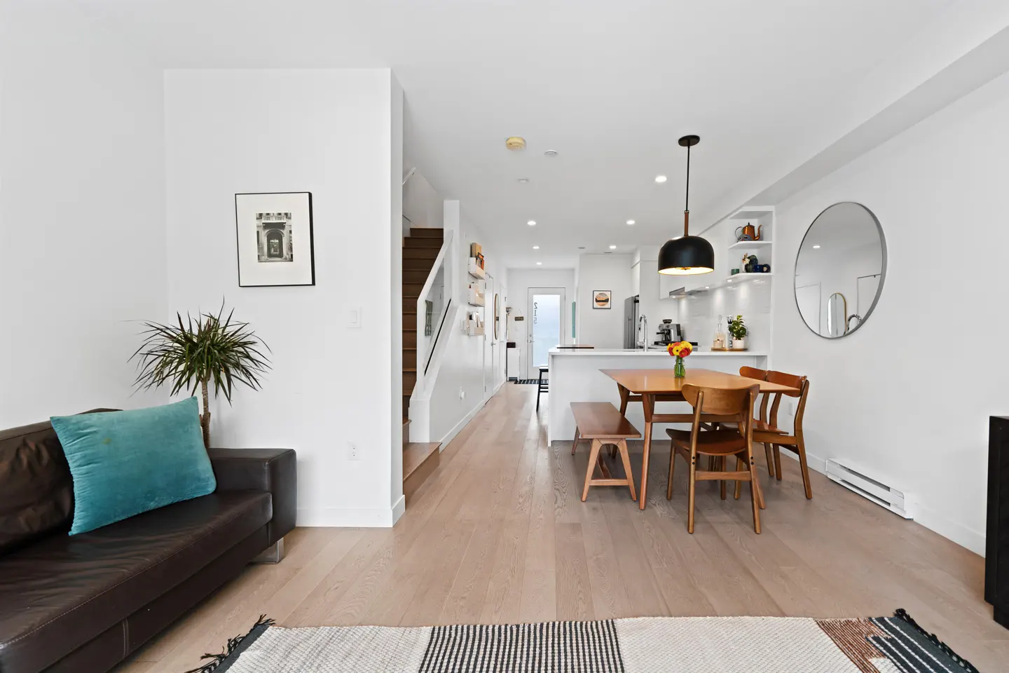 Bright, modern home interior with a brown leather sofa, wood dining table, and white kitchen. Stairs lead to the upper level.