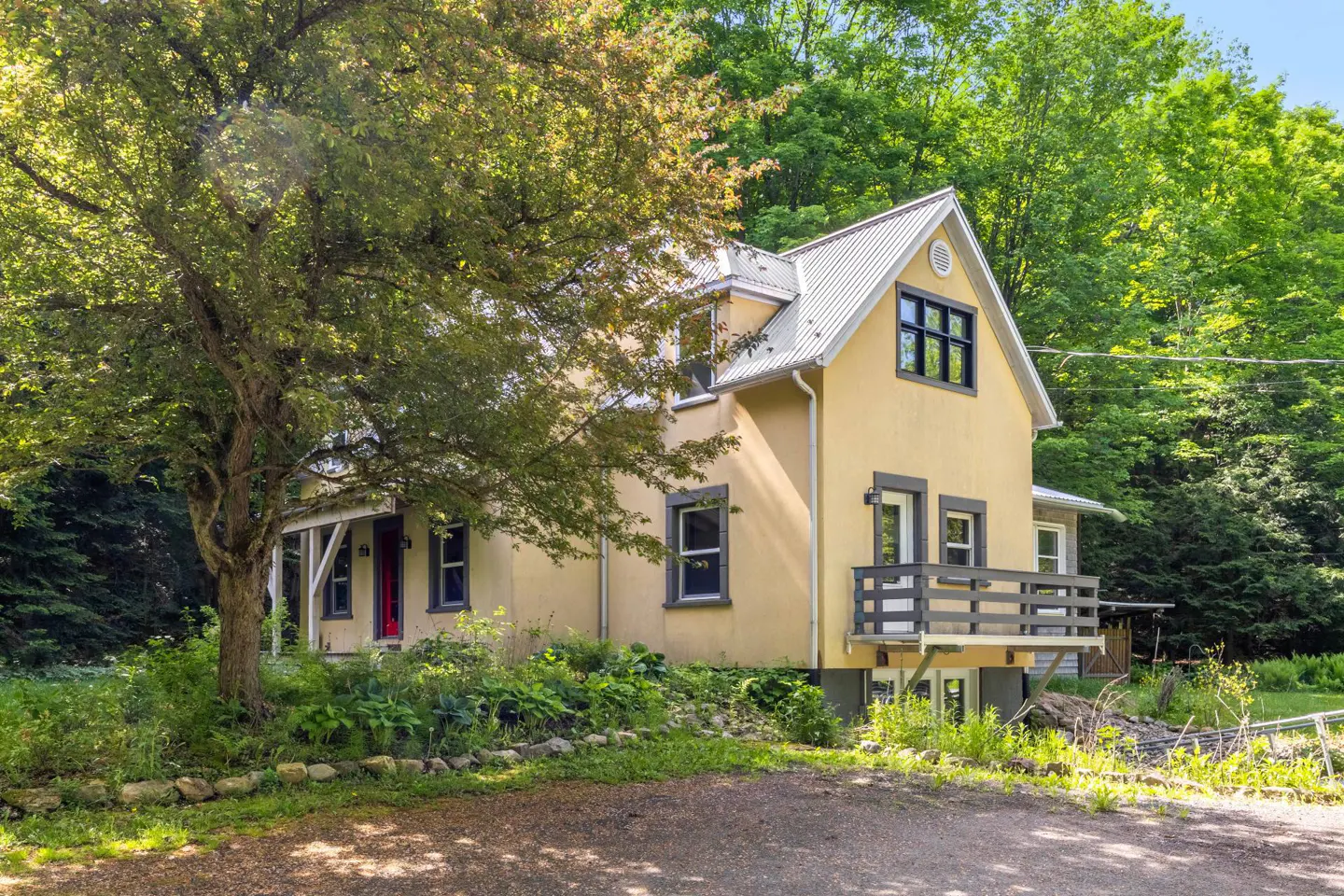 Yellow two-story house with a metal roof, wooden balcony, and dark trim, nestled in a lush, tree-filled forest setting.