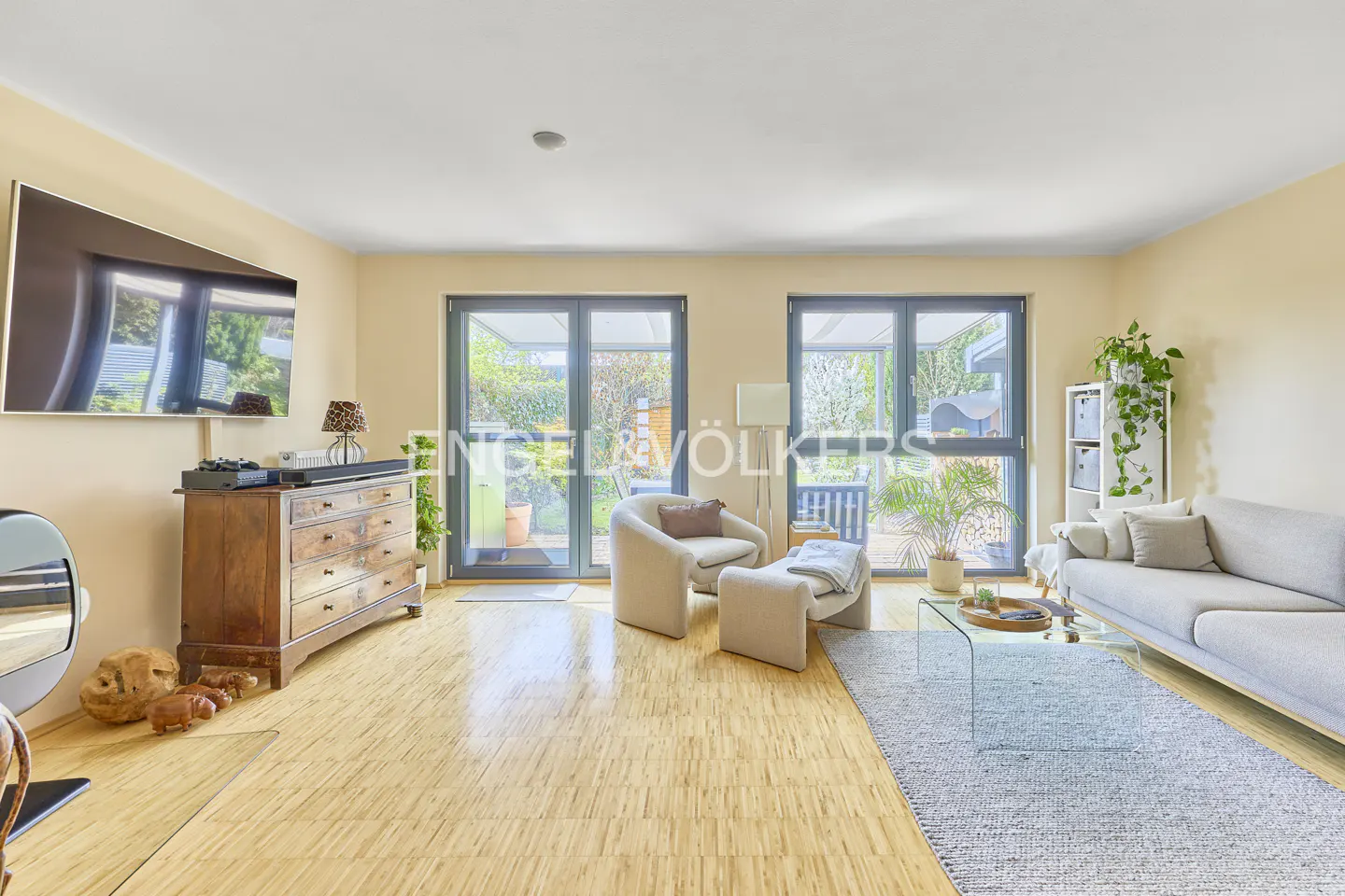 Living room with light wood floors, a gray sofa, and a glass coffee table on a gray rug. Two sets of glass doors lead to a garden.