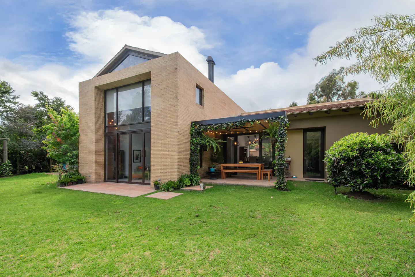 Exterior view of a modern two-story house with a green lawn, a patio with a wooden table, and a pergola covered in vines.