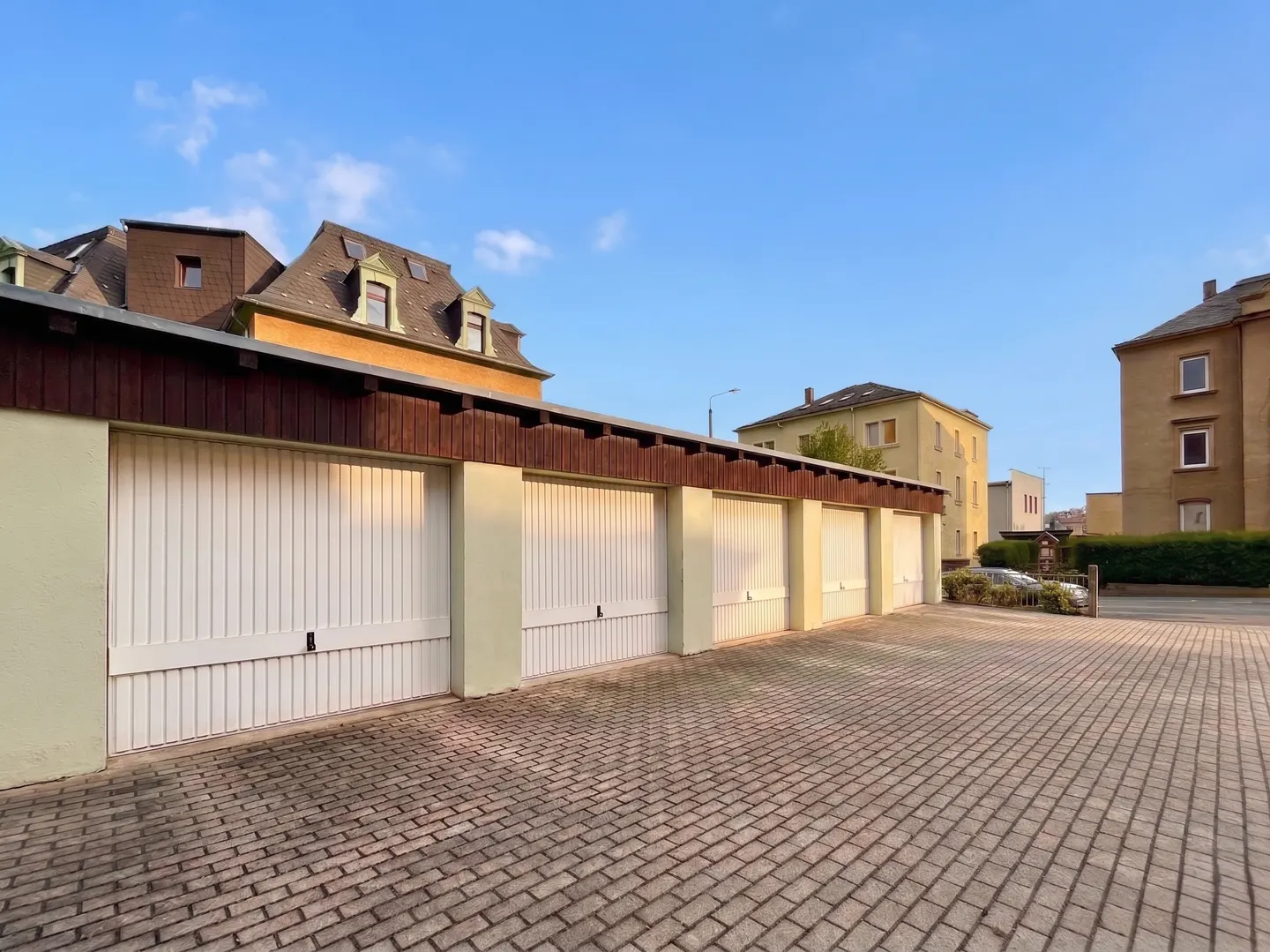 Row of white garages with brown trim under a blue sky, brick driveway, and apartment buildings in the background.