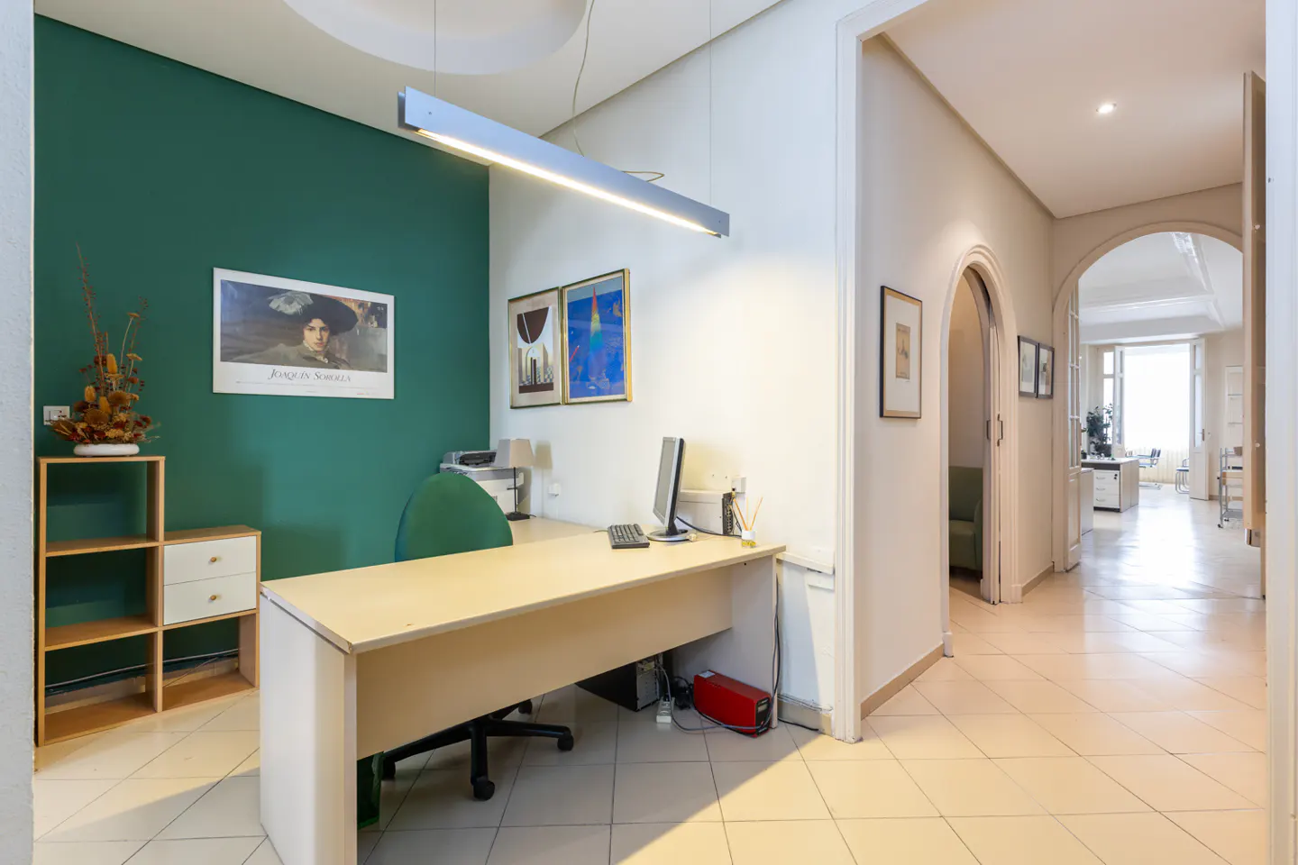 Office space with a desk, computer, and green accent wall. Hallway with arched doorways leads to a bright, distant office.