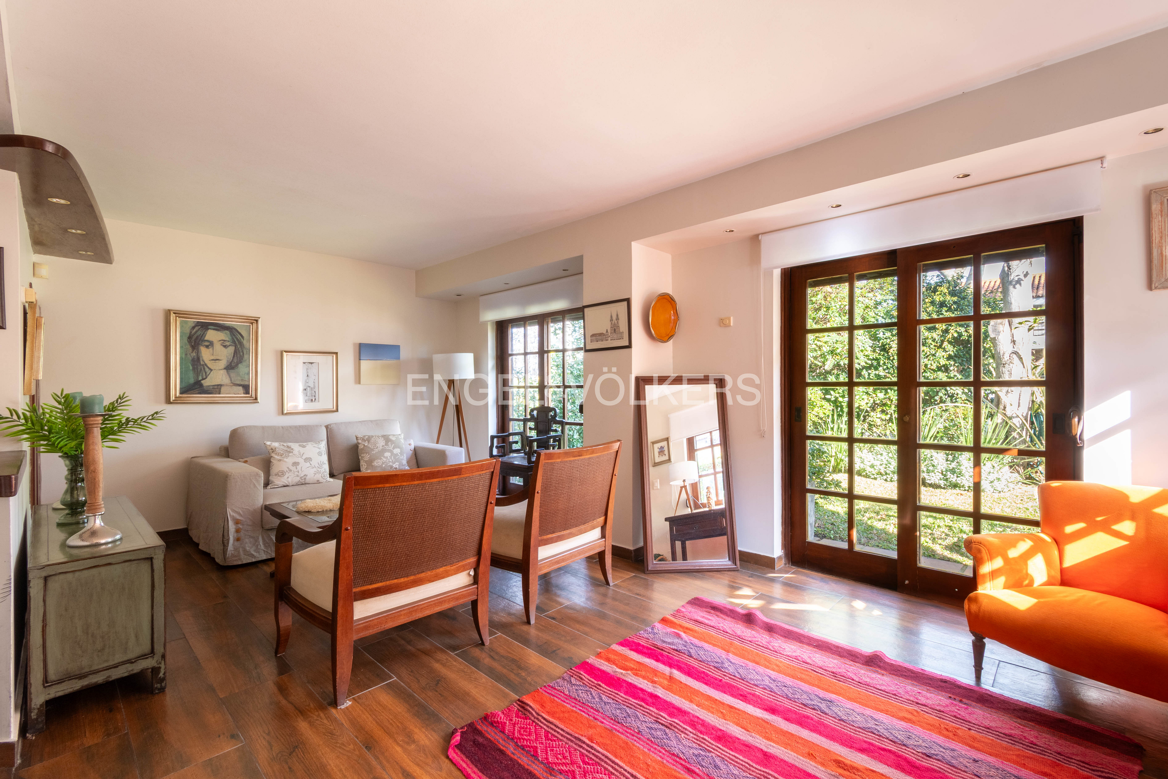 Living room with a beige sofa, two wooden armchairs, a bright orange armchair, and a striped rug on a wooden floor.