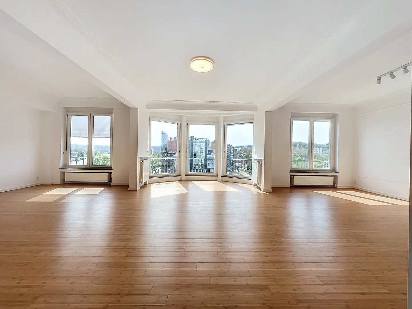 Bright, empty room with hardwood floors, white walls, and large windows offering a city view.