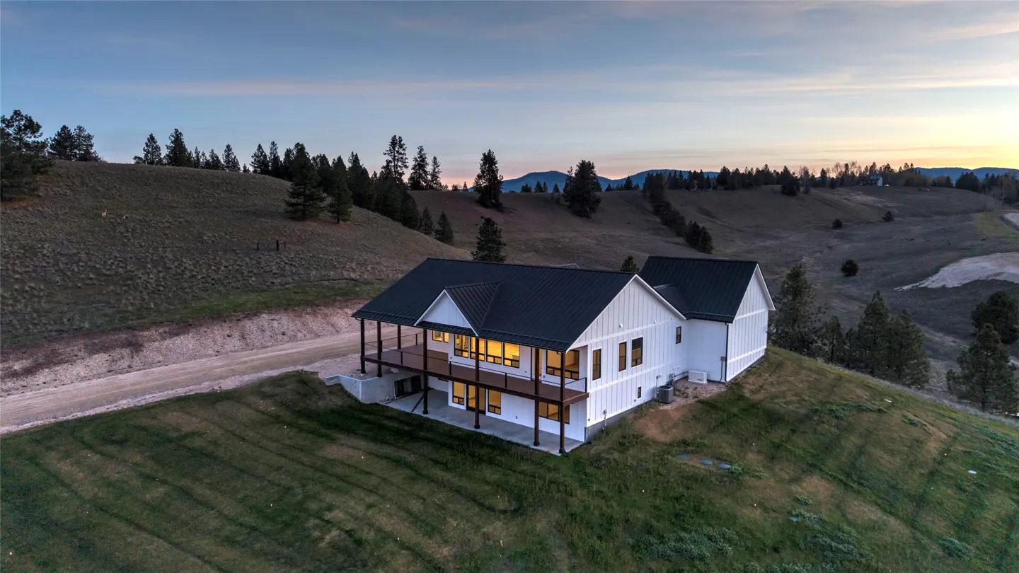 Aerial view of a modern white farmhouse with a black roof on a grassy hill at dusk. Trees and mountains are in the background.