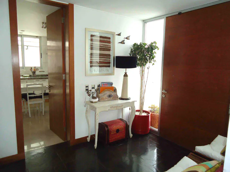 A bright room with a dark wood floor features a white table with a lamp, art, and a plant near a large window and a door.