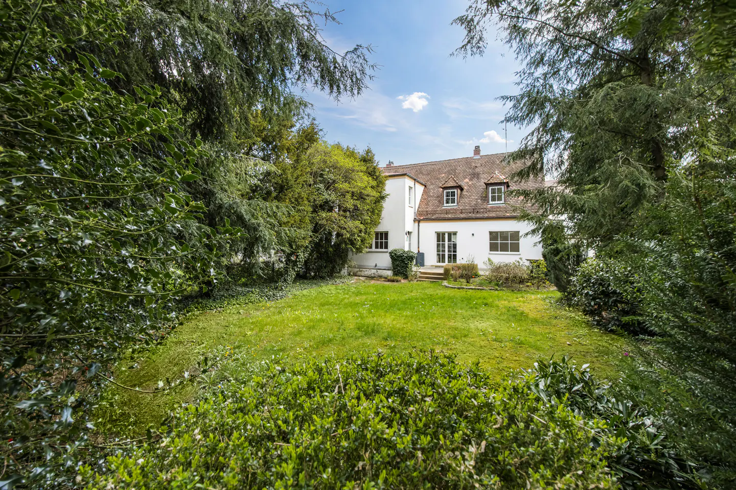 View of a white house with a brown roof, surrounded by green trees and a lawn.