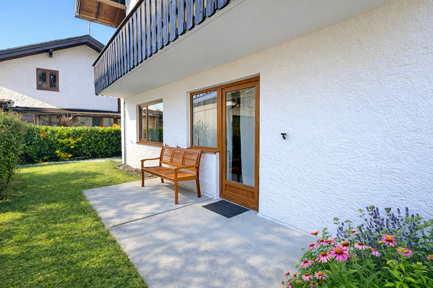 Exterior view of a white building with a wooden bench on a concrete patio, next to a lawn and flower bed. A wooden door and window are visible.