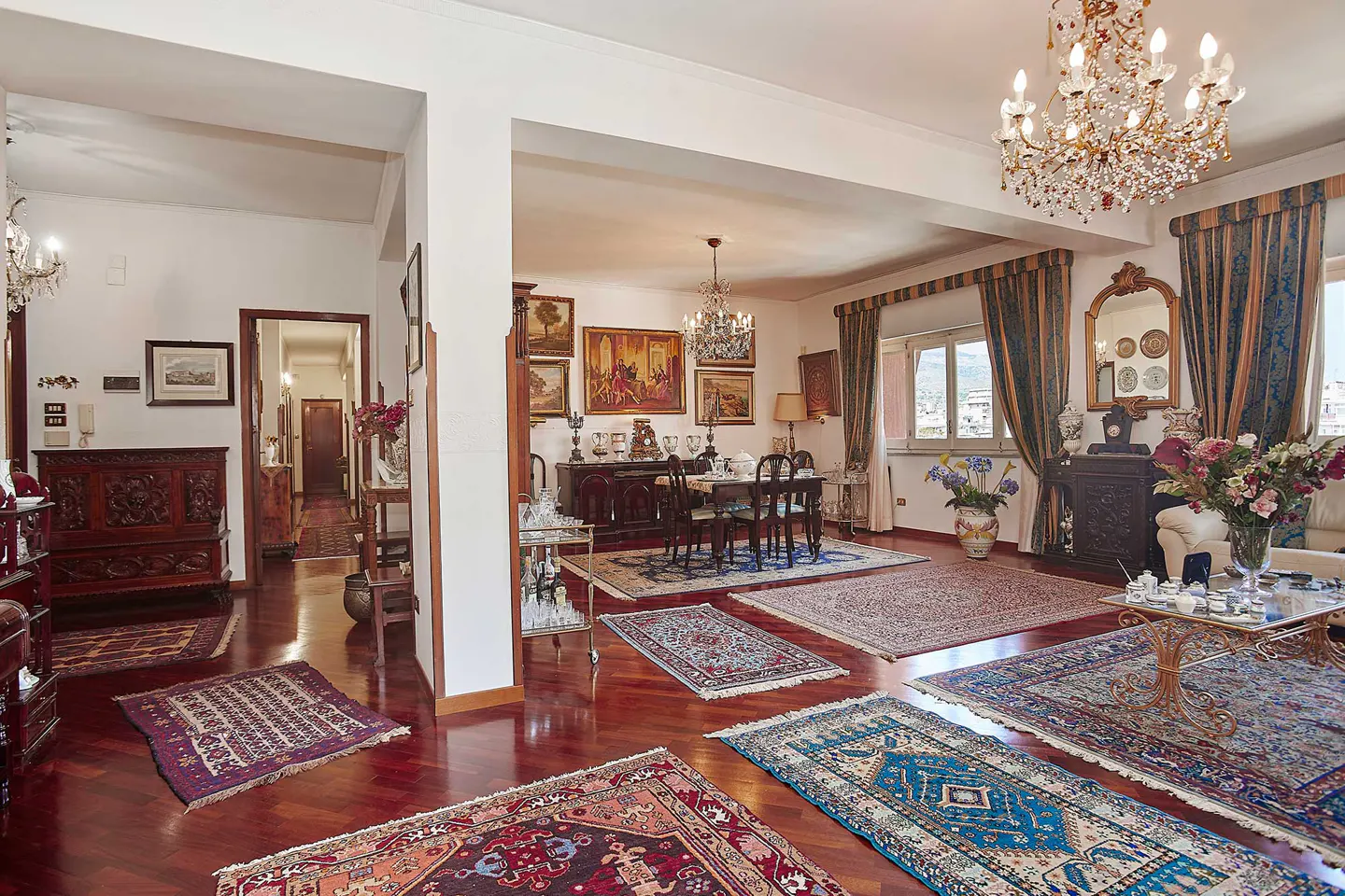 A spacious living room with hardwood floors and multiple Persian rugs. A dining table, chandelier, and various antique furnishings are visible.