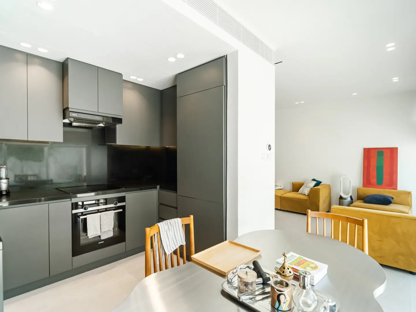 Open-concept kitchen and living room with gray cabinets, stainless steel table, and yellow sofas.