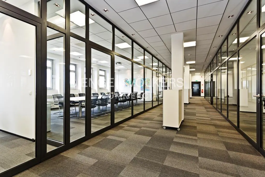 Office hallway with glass-walled rooms. A conference room with a table and chairs is visible. The floor is carpeted in gray and brown.
