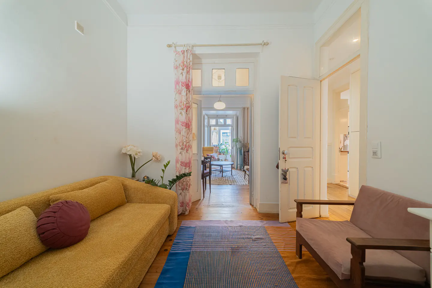 Bright living room with a yellow sofa, pink chair, and a blue striped rug on a wooden floor. A floral curtain hangs near an open doorway.