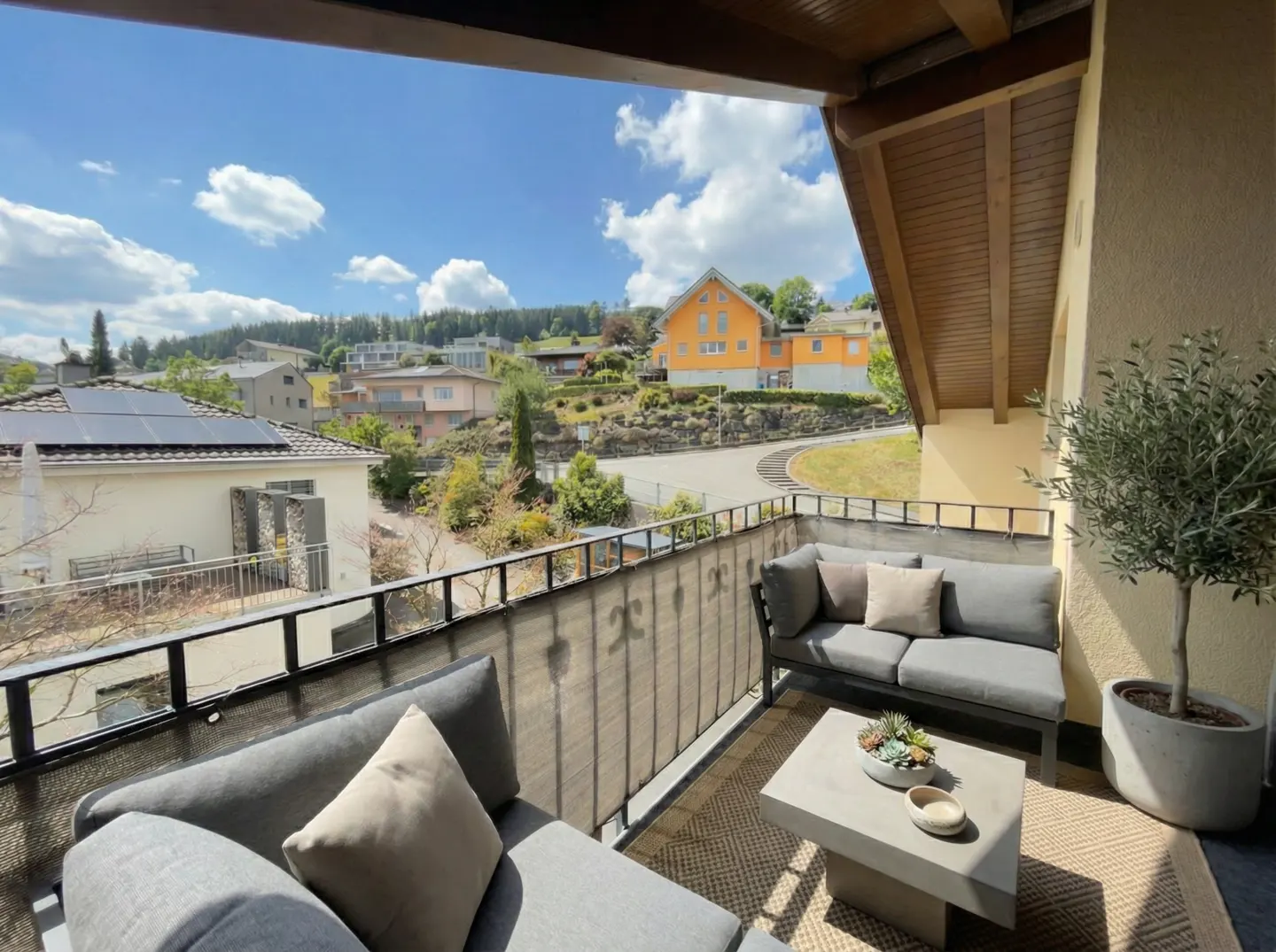 Balcony view with gray sofas, a table, and potted tree. Outside, houses dot a green hillside under a blue sky with white clouds.