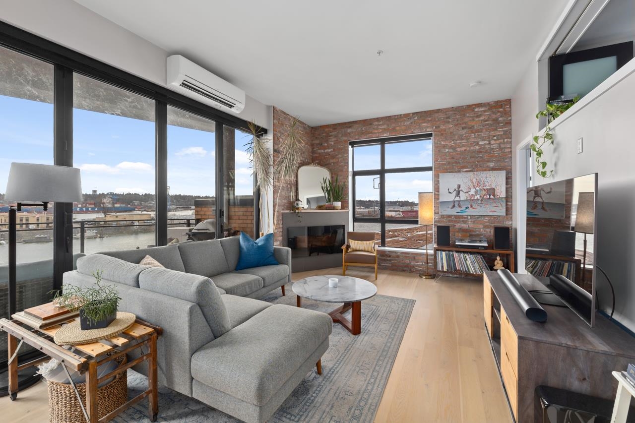 Bright living room with a gray sectional sofa, brick wall, fireplace, and large windows overlooking a city and water view.