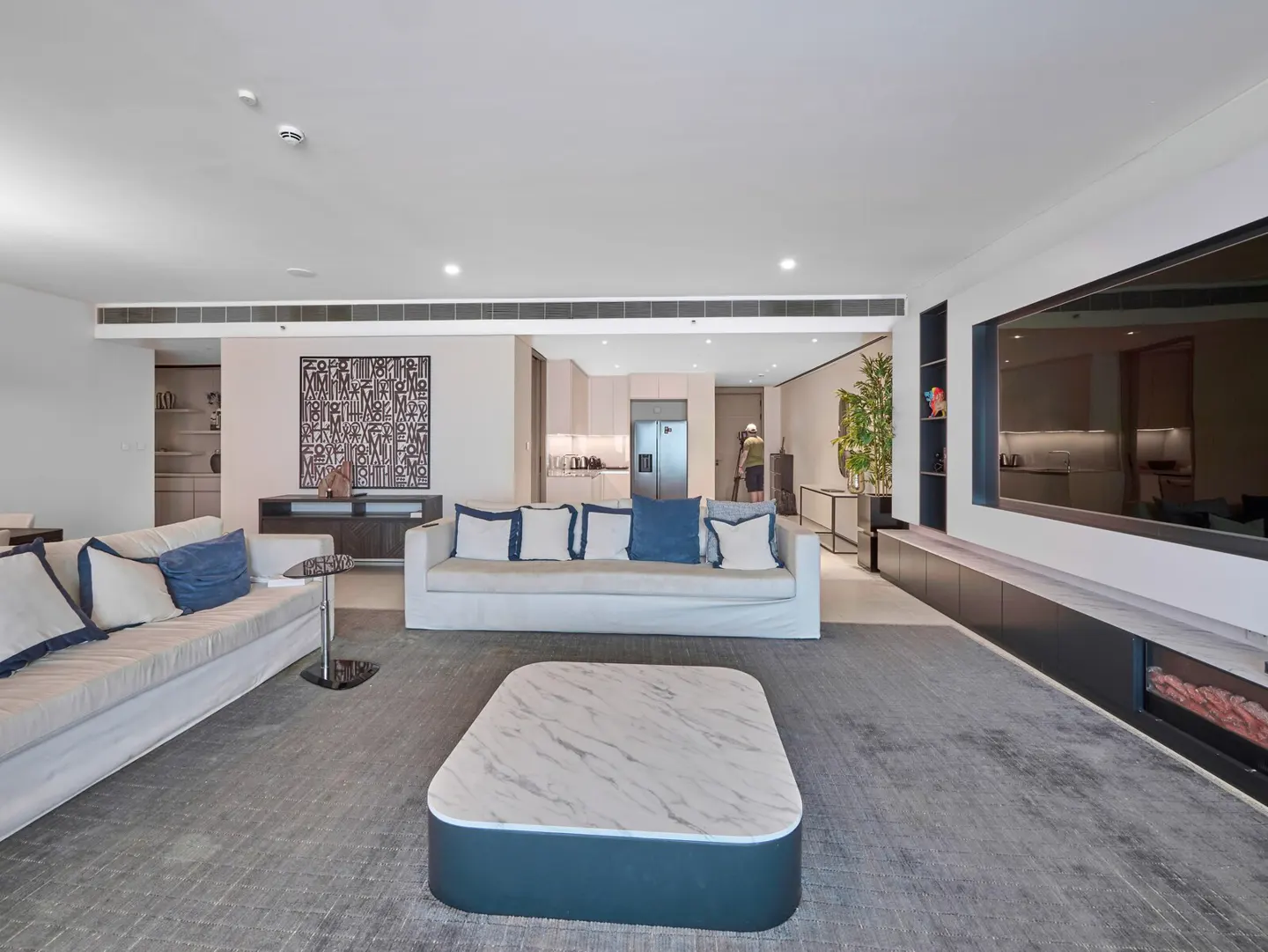 A modern living room with white sofas, blue pillows, and a marble coffee table on a gray rug. A large TV is mounted on the wall.